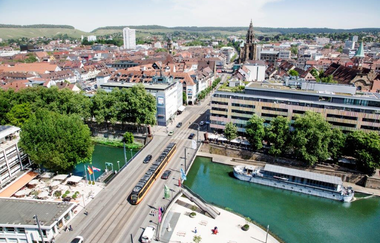 Das Bild zeigt  ein Luftbild von Heilbronn. Eine S-Bahn fährt über die Friedrich-Ebert-Brücke. Unter der Brücke fließt das türkis-grüne Wasser des Neckars. Die Blätter der Bäume sind grün. Im Hintergrund sieht man die Gebäude in der Innenstadt.