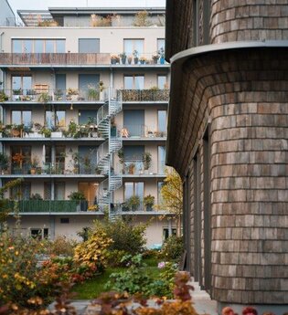 Auf dem Bild sind mehrere Stockwerke eines modernen Gebäudes mit Balkonen und einer Wendeltreppe zu sehen.  Es gibt einen begrünten Innenhof mit herbstlichen Pflanzen und Bäumen. Ein weiteres Gebäude ist auf der linken Seite des Bildes.