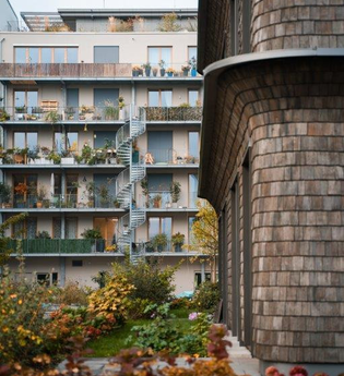 Auf dem Bild sind mehrere Stockwerke eines modernen Gebäudes mit Balkonen und einer Wendeltreppe zu sehen.  Es gibt einen begrünten Innenhof mit herbstlichen Pflanzen und Bäumen. Ein weiteres Gebäude ist auf der linken Seite des Bildes.