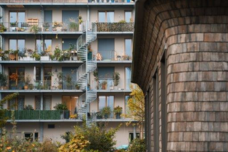 Auf dem Bild sind mehrere Stockwerke eines modernen Gebäudes mit Balkonen und einer Wendeltreppe zu sehen.  Es gibt einen begrünten Innenhof mit herbstlichen Pflanzen und Bäumen. Ein weiteres Gebäude ist auf der linken Seite des Bildes.