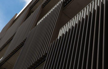Auf dem Bild ist ein Gebäude von unten entlang der Fassade fotografiert. An der Fassade sind viele einzelne Stäbe. Das Gebäude hat viele  Loggien.