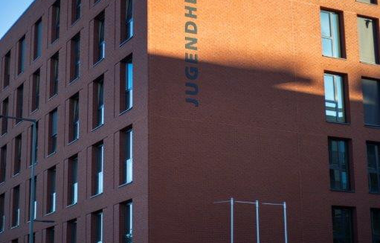 Das Foto zeigt ein großes Gebäude aus roten Backsteinen. Darauf ist die Aufschrift "Jugendherberge" in dunklen Großbuchstaben. Ein Teil der Fassade ist von der Sonne beschienen, der Rest liegt im Schatten. Der Himmel ist wolkenlos.