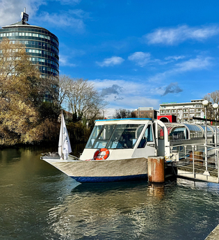 Das Bild zeigt ein Ausflugsboot an der Anlegerstelle "Marrahaus". Ein hohes, modernes Gebäude und Bäume sind im Hintergrund zu sehen. Der Himmel strahlt blau, fast wolkenlos. Die Stimmung ist friedlich und einladend.