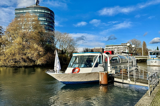 Das Bild zeigt ein Ausflugsboot an der Anlegerstelle "Marrahaus". Ein hohes, modernes Gebäude und Bäume sind im Hintergrund zu sehen. Der Himmel strahlt blau, fast wolkenlos. Die Stimmung ist friedlich und einladend.