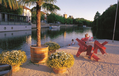 Das Bild zeigt einen Fluss gesäumt durch einen Sandstrand auf dem drei Blumenkübel buschig bepflanzt und eine Palme in einem Fass eingepflanzt. Im Hintergrund spielt ein Kind am Wasser. Erwachsene schauen zu. Auf der gegenüberliegenden Seite des Flusses i