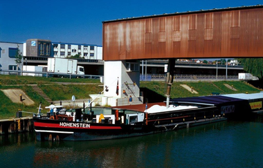 Auf dem Bild ist ein Schiff zu sehen das in verschiedenen Kammer mit Salz befüllt wird. Im Hintergrund erkennt man Bürogebäude und einen weißen Lastkraftwagen