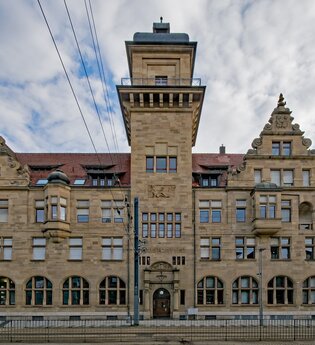 Das Bild zeigt ein großes, imposantes Gebäude aus Sandstein mit einer Art Turm in der Mitte. Das Gebäude hat eine symmetrische Fassade mit vielen Fenstern und dekorativen Elementen wie Giebeln und Wappen. Der Himmel ist bewölkt.