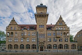 Das Bild zeigt ein großes, imposantes Gebäude aus Sandstein mit einer Art Turm in der Mitte. Das Gebäude hat eine symmetrische Fassade mit vielen Fenstern und dekorativen Elementen wie Giebeln und Wappen. Der Himmel ist bewölkt.