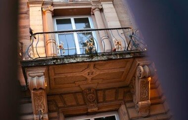 Das Bild zeigt eine Nahaufnahme eines Gebäudes. Zu sehen sind drei Etagen mit 3 Fenstern, die jeweils von architektonischen Elementen wie Säulen, Zierleisten und Gesimsen umrahmt sind. In der Mitte befindet sich ein Balkon mit verziertem Eisengeländer.