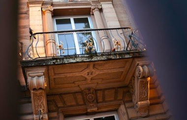 Das Bild zeigt eine Nahaufnahme eines Gebäudes. Zu sehen sind drei Etagen mit 3 Fenstern, die jeweils von architektonischen Elementen wie Säulen, Zierleisten und Gesimsen umrahmt sind. In der Mitte befindet sich ein Balkon mit verziertem Eisengeländer.