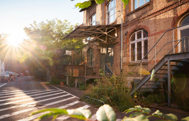 Das Bild zeigt ein Backsteingebäude mit einer Eisentreppe, die zu einem kleinen führt. Die Sonne scheint hell und wirft lange Schatten auf die Straße. Am Straßenrand sind Tische und Stühle aufgestellt. Im Vordergrund sind unscharfe Blätter zu sehen.