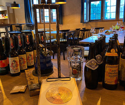 Auf einem Biertresen stehen viele verschiedene Bierflaschen. Im Vordergrund liegt ein Klemmbrett mit einem Block auf dem eine Scheibe zu sehen ist. Im Hintergrund sieht man die Räumlichkeiten, große mit Butzenglas verglaste Fenster, Tische  und Stühle