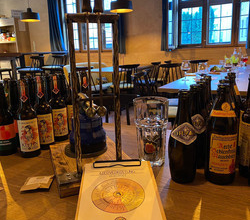 Auf einem Biertresen stehen viele verschiedene Bierflaschen. Im Vordergrund liegt ein Klemmbrett mit einem Block auf dem eine Scheibe zu sehen ist. Im Hintergrund sieht man die Räumlichkeiten, große mit Butzenglas verglaste Fenster, Tische  und Stühle