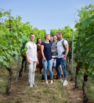 In der Mitte des Bildes steht eine Gruppe aus Personen, alle mit einem breiten Lächeln. Sie stehen zwischen zwei Reben. Die Blätter sind grün, der Himmel ist blau und fast wolkenlos. Die Stimmung wirkt locker und fröhlich.
