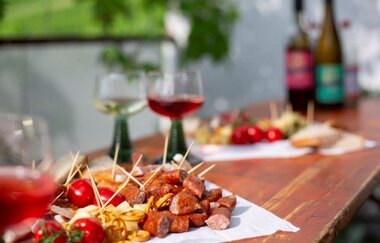 Im Fokus des Bildes sieht man Snacks auf einem Biertisch. Die Tomaten, Käsewürfel, Wurststückchen und Salzbrezeln liegen auf einer Serviette mit Zahnstochern in sich. Im Hintergrund stehen zwei Gläser mit Rot- und Weißwein, sowie weitere Snacks und Wein.