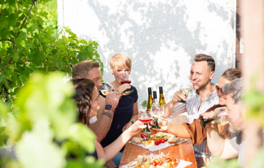 Auf dem Bild sieht man eine Gruppe am Holztisch im Freien. Jeder trinkt aus seinem Glas ein Schluck Wein. Die Szene ist von grünen Blättern an Ästen und strahlendem Sonnenlichtumgeben, was eine entspannte Atmosphäre schafft.