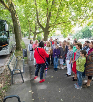 Auf dem Bild ist eine Gruppe von Touristen zu sehen, die sich vor einem Reisebus versammelt hat. Eine Person in rotem Outfit erklärt etwas, während die anderen aufmerksam zuhören. Der Straßenrand ist von grünen Bäumen umgeben, dahinter fließt der Neckar.