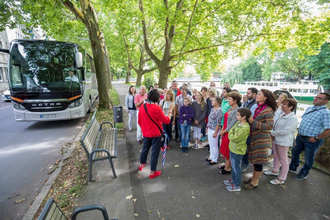 Auf dem Bild ist eine Gruppe von Touristen zu sehen, die sich vor einem Reisebus versammelt hat. Eine Person in rotem Outfit erklärt etwas, während die anderen aufmerksam zuhören. Der Straßenrand ist von grünen Bäumen umgeben, dahinter fließt der Neckar.