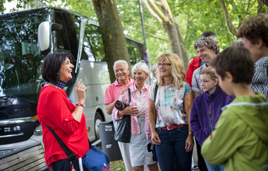 Das Bild zeigt eine Dame in einem rotem Shirt, die einer Gruppe etwas erklärt. Die Gruppe hört aufmerksam zu. Im Hintergrund ist ein silberner Reisebus zu sehen, der unter den grünen Bäumen am Straßenrand geparkt ist.
