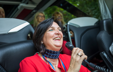 Auf dem Bild ist eine Dame zu sehen, die auf einem dunklen Bussitz sitzt und ein Mikrofon in der Hand hält. Im Hintergrund kann man drei Menschen sehen, die aus dem Fenster schauend beobachten und zuhören.