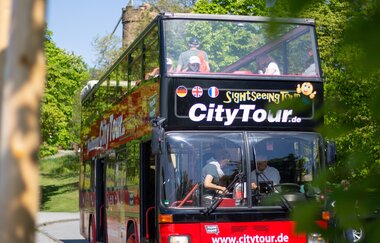 Roter Doppeldeckerbus mit offenem Dach aufgenommen von schräg links vorne. Die Sonne scheint und der Bus ist von grünen Bäumen umgeben | © Heilbronn Marketing GmbH