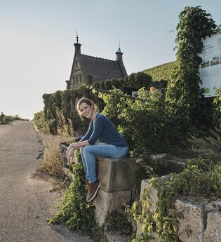 Eine Frau in Jeans sitzt entspannt auf einer Steinmauer entlang eines Weges, umgeben von grünen Weinreben. Im Hintergrund steht ein kleines Gebäude mit einem spitzen Dach und zwei Spitzen hinten und vorne. Die Szene wird von warmem Sonnenlicht beleuchtet.