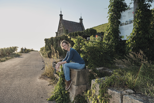 Eine Frau in Jeans sitzt entspannt auf einer Steinmauer entlang eines Weges, umgeben von grünen Weinreben. Im Hintergrund steht ein kleines Gebäude mit einem spitzen Dach und zwei Spitzen hinten und vorne. Die Szene wird von warmem Sonnenlicht beleuchtet.