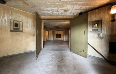 Das Foto zeigt einen verlassenen Innenraum aus Beton. Verwitterte Wände und schwere, olivgrüne Türen schaffen eine Atmospäre von Verlassenheit und Kälte. Der Betonboden ist abgenutzt, der Raum wirkt leer und still.