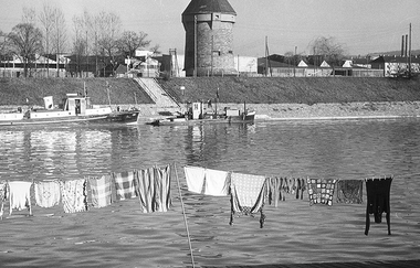 Das Bild zeigt eine historische schwarz-weiß Aufnahme. Im Vordergrund ist eine Wäscheleine am Ufer des Neckars. Hinter dem Neckar sieht  man das andere Ufer mit einem kleinen Schiff auf der linken Seite und dem Theresienturm auf der rechten Seite.