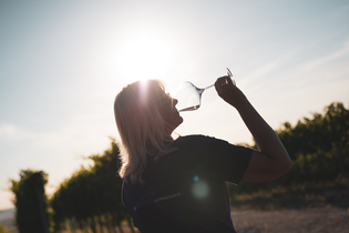 Ein Frau mit längeren, blonden Haaren die ein Glas Weißwein trinkt im Gegenlicht aufgenommen. Schöne Lichtstimmung im Sonnenuntergang mit ein Weinbergen im Hintergrund. | © Nicole Halter