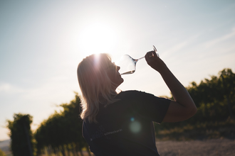 Ein Frau mit längeren, blonden Haaren die ein Glas Weißwein trinkt im Gegenlicht aufgenommen. Schöne Lichtstimmung im Sonnenuntergang mit ein Weinbergen im Hintergrund. | © Nicole Halter