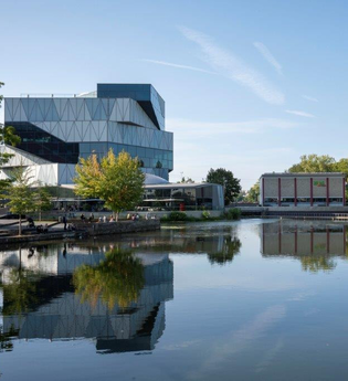 Der Neckar im Vordergrund. Im Wasser spiegelt sich die experimenta. Das Ufer ist von Büschen und Bäumen gesäumt