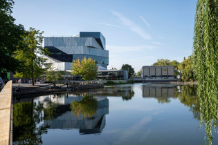Der Neckar im Vordergrund. Im Wasser spiegelt sich die experimenta. Das Ufer ist von Büschen und Bäumen gesäumt