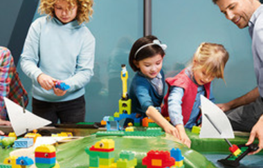 Im Bild vorne sind bunte Gebilde aus großen Lego-Steinen zu sehen die auf große grüne Lego Unterplatten fantasievoll gestaltet wurden. Hinter dem Gebilde sind drei Kinder und ein Mann zu sehen  die fleißig weiterbauen