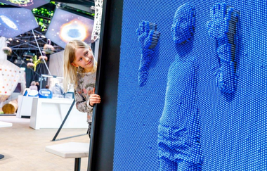 rechts im Bild ist eine große blaue, begehbare Nadelwand zu erkennen das die Umrisse eines kleinen Mädchens zeigt. Das Mädchen, das sich gegen die Stiftwand gelehnt hatte, und deren Umrisse zu sehen sind schaut gespannt auf das entstandene Bild