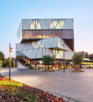 Moderner Bau mit dreieckigen Fassadenelementen und großen Glasflächen. Daneben ist ein Platz mit Wiesen und Beeten und ein gepflasterter Platz. Das Foto ist in der Dämmerung aufgenommen.