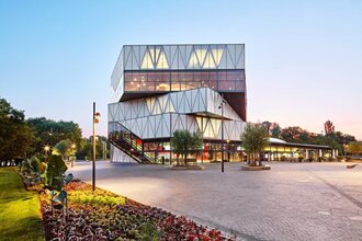 Moderner Bau mit dreieckigen Fassadenelementen und großen Glasflächen. Daneben ist ein Platz mit Wiesen und Beeten und ein gepflasterter Platz. Das Foto ist in der Dämmerung aufgenommen.