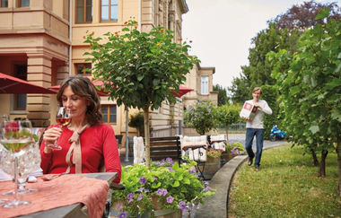 In der linken Hälfte sitzt eine Frau mit einem Weinglas in der Hand an einem gedeckten Tisch. In der rechten Hälfte läuft eine Person auf einer Gras bewachsenen Fläche, zwischen kleinen Bäumen, mit einem Weinkarton in der Hand.