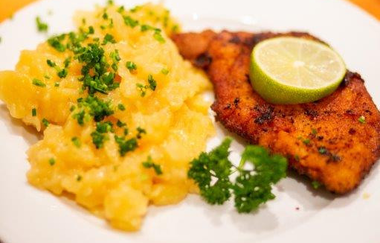 Fackel-Weinwanderung | © Auf dem Bild ist eine weiße Serviette zu sehen auf der Messer und Gabel liegen. Darüber steht ein weißer, runder Teller auf dem Kartoffelsalat, garniert mit kleingehackter Petersilie und einem panierten Schnitzel mit Limette garniert zu sehen ist