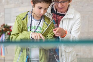 Das Bild zeigt einen Jungen und eine ältere Dame, die auf ein weißes Modell einer Stadt schauen. Das Modell befindet sich unter einem Glaskasten.