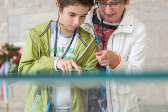 Das Bild zeigt einen Jungen und eine ältere Dame, die auf ein weißes Modell einer Stadt schauen. Das Modell befindet sich unter einem Glaskasten.