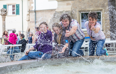 Das Bild zeigt eine Familie mit zwei Kindern, die auf einem Brunnen sitzen und knien und mit den Händen Wasser in Richtung der Linse spritzen. Die Familie wirkt amüsiert.