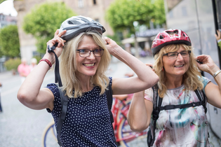 Das Bild zeigt zwei lachende Damen, die sich einen Helm aufsetzen. Im Hintergrund sieht man Fahrräder, einen gepflasterten Platz, grüne Bäume und Gebäude.