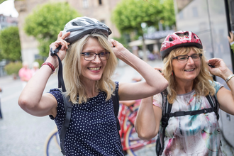 Das Bild zeigt zwei lachende Damen, die sich einen Helm aufsetzen. Im Hintergrund sieht man Fahrräder, einen gepflasterten Platz, grüne Bäume und Gebäude.