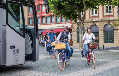 Das Bild zeigt 4 Personen die hintereinander auf Fahrrädern fahren. Links steht ein Bus mit offener Türe. Im Hintergrund sieht man ein rotes und ein bräunliches Gebäude. Die Gruppe wirkt locker und amüsiert.