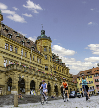 Blick auf den Marktplatz mit Rathaus in Rothenburg o.d.T.  Hier fahren gerade drei Radelnde über das Kopfsteinpflaster.