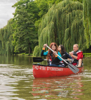 Das Bild zeigt 3 Personen in einem roten Kanu mit Paddeln in der Hand. Am Ufer sind grüne Wiesen und Trauerweiden sowie andere Bäume. Der Himmel ist bewölkt.