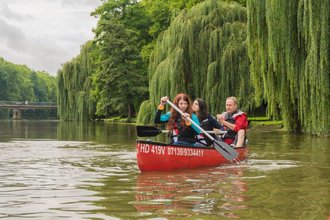 Das Bild zeigt 3 Personen in einem roten Kanu mit Paddeln in der Hand. Am Ufer sind grüne Wiesen und Trauerweiden sowie andere Bäume. Der Himmel ist bewölkt.