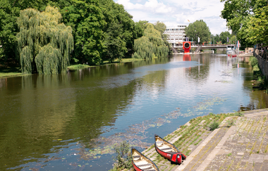 Das Bild zeigt vier rote 3 Personen Kanus an einem gepflastertem Einstieg in den Neckar. Auf der anderen Seite des Ufers sind grüne Bäume und Wiesen. Die Sonne scheint hell und der Himmel ist fast wolkenlos.