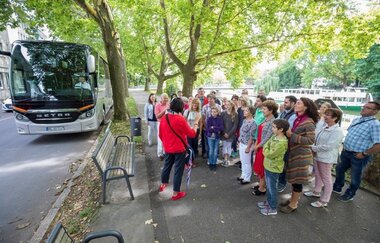 im Bild links sieht man einen geparkten Bus mit der Front nach vorne. Rechts davor stehen Teilnehmer der Busreise und lauschen den Worten der Stadtführerin. Im Hintergrund ist der Neckar mit einem Schiff erkennbar